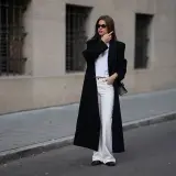Elegancka kobieta w czarnym płaszczu i białych, szerokich spodniach. Idealne buty do szerokich spodni to te, które dodają stylizacji szyku, jak te czarne mokasyny.
